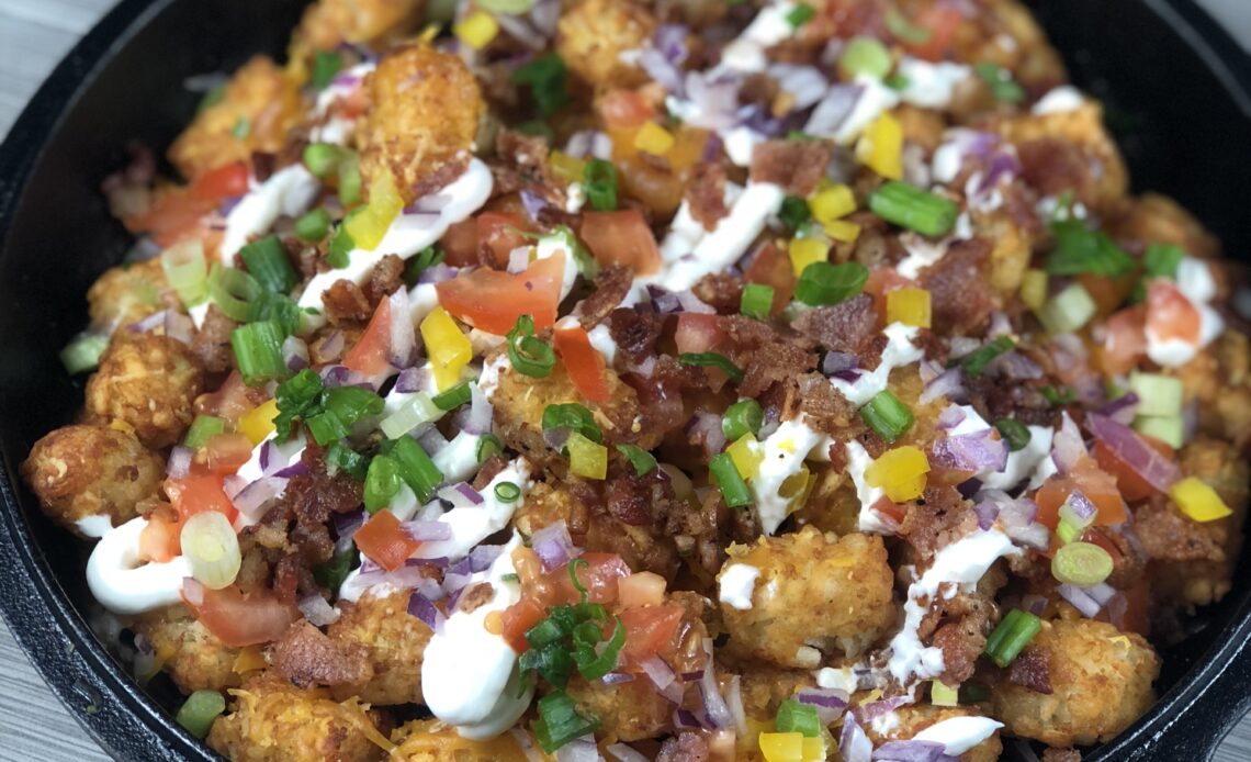 A photo of a cast iron skillet filledwith tater tots topped with melted cheese, sour cream, peppers, onions, tomatoes and scallion.