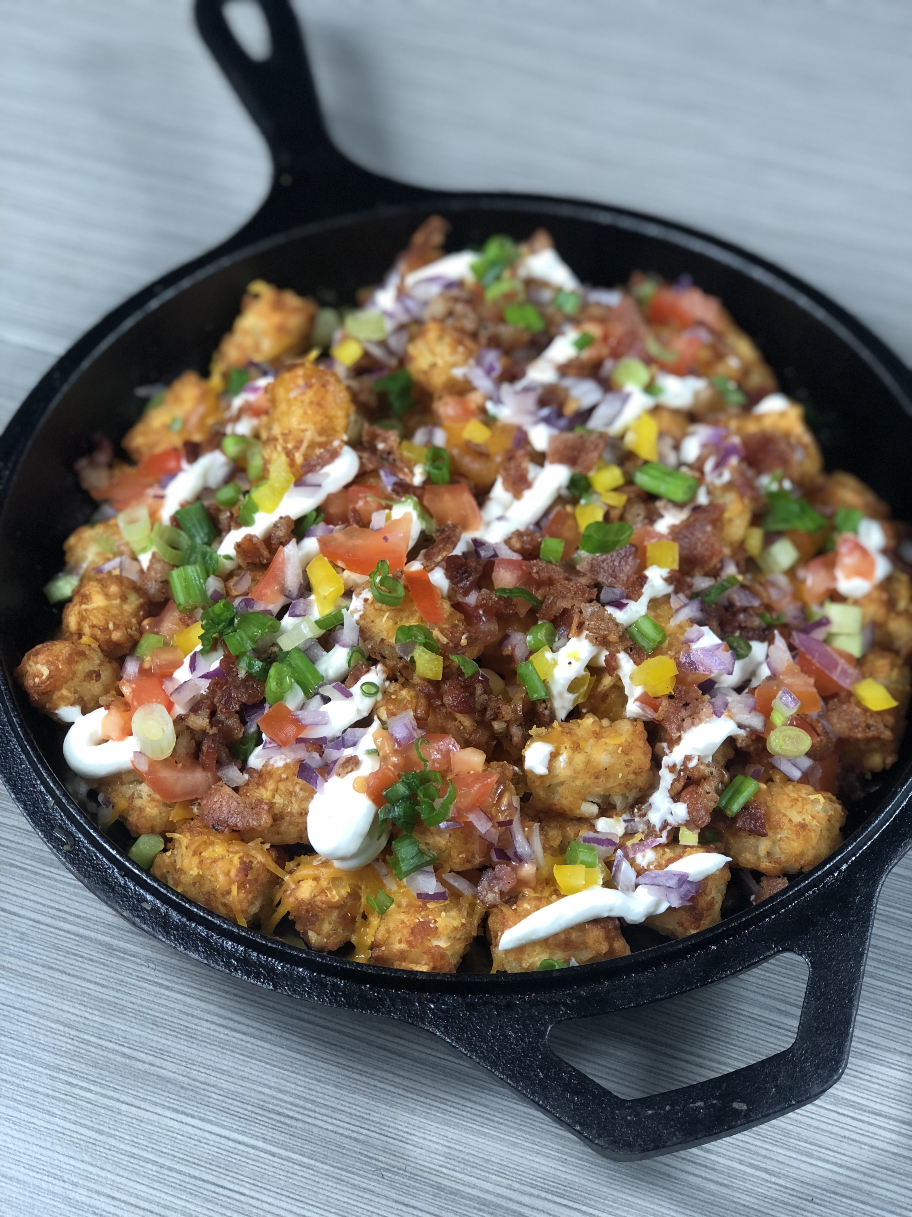 A photo of a cast iron skillet filledwith tater tots topped with melted cheese, sour cream, peppers, onions, tomatoes and scallion.