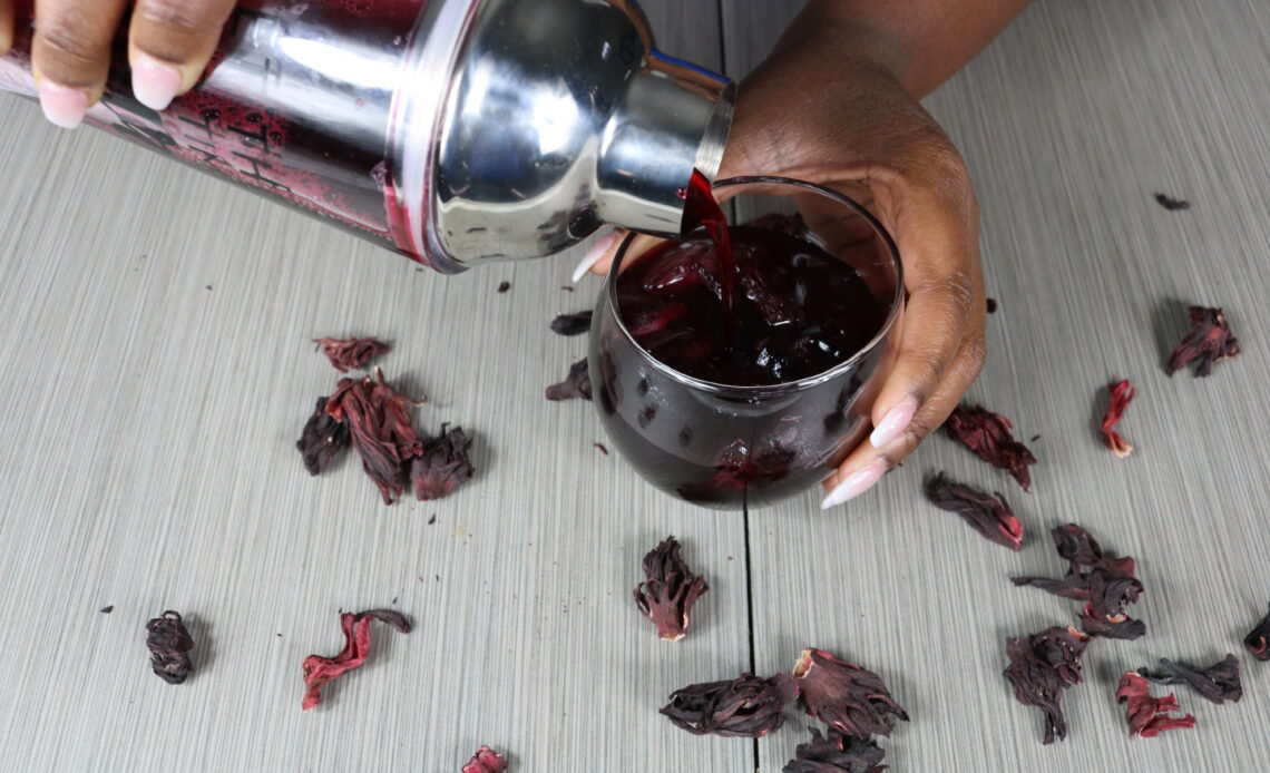 A glass of ice with sorrel being poured from a drink shaker surrounded by dried hibiscus in the background.
