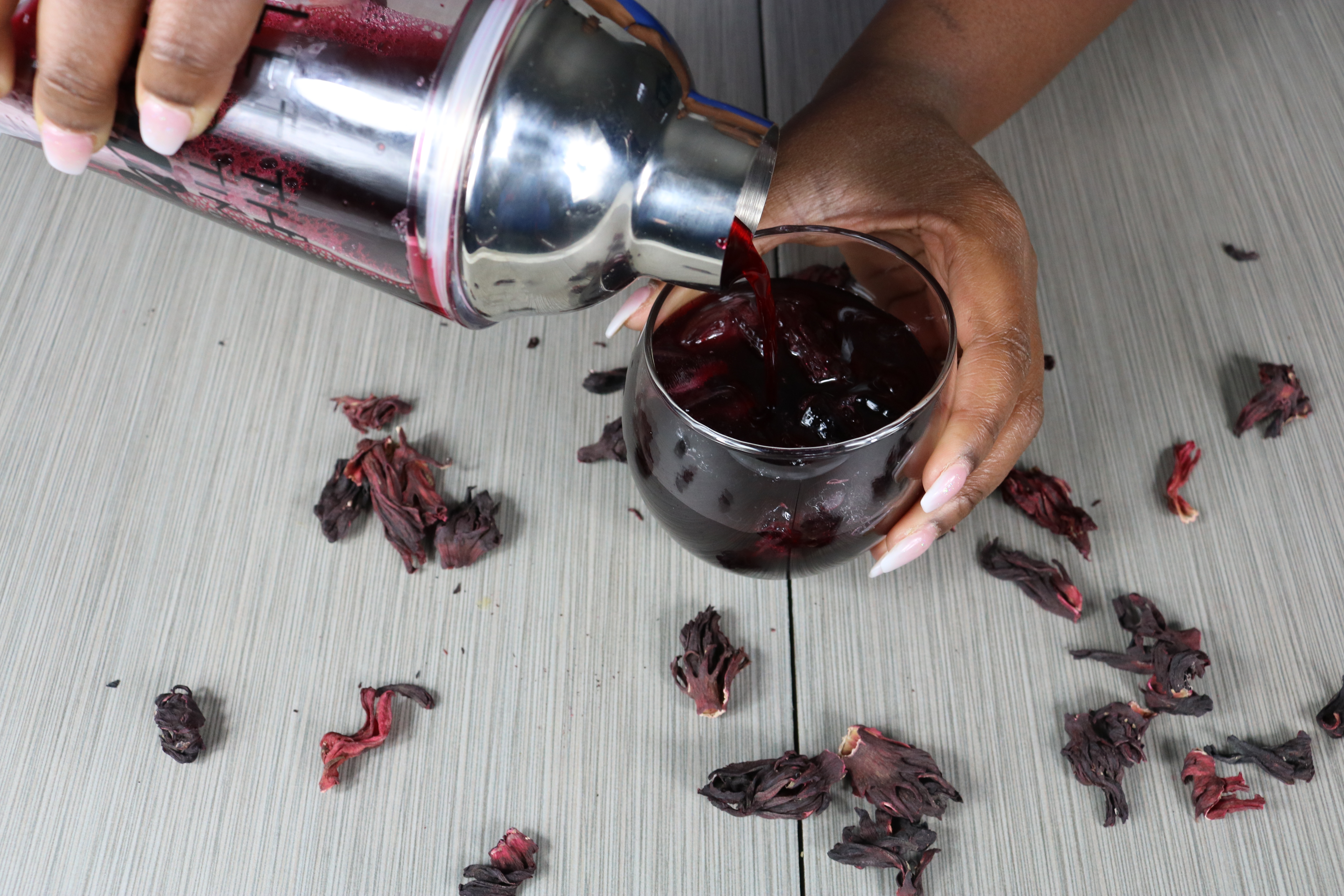 A glass of ice with sorrel being poured from a drink shaker surrounded by dried hibiscus in the background.