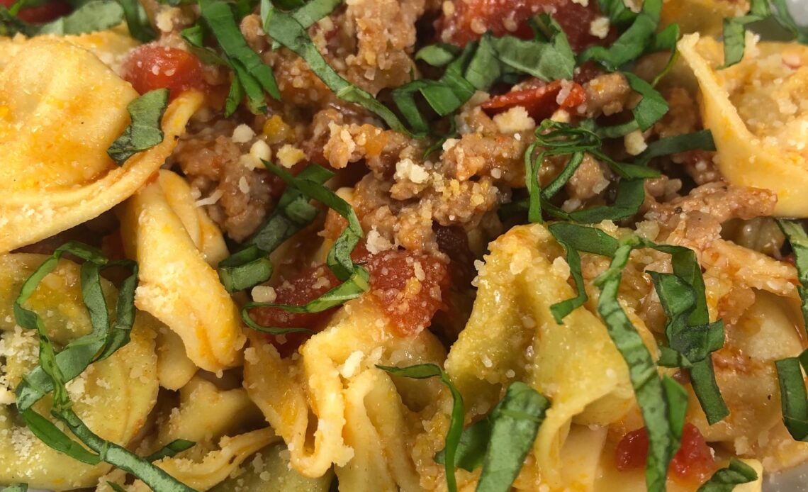 A bowl of tortellini topped with italian sausage, diced tomatoes, grated parmesan and strips of fresh basil.