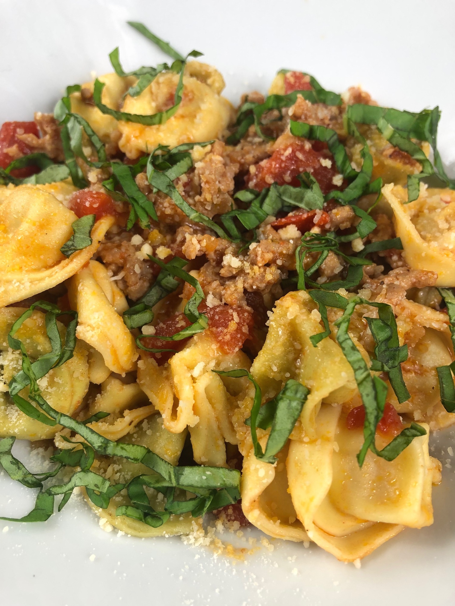 A bowl of tortellini topped with italian sausage, diced tomatoes, grated parmesan and strips of fresh basil.