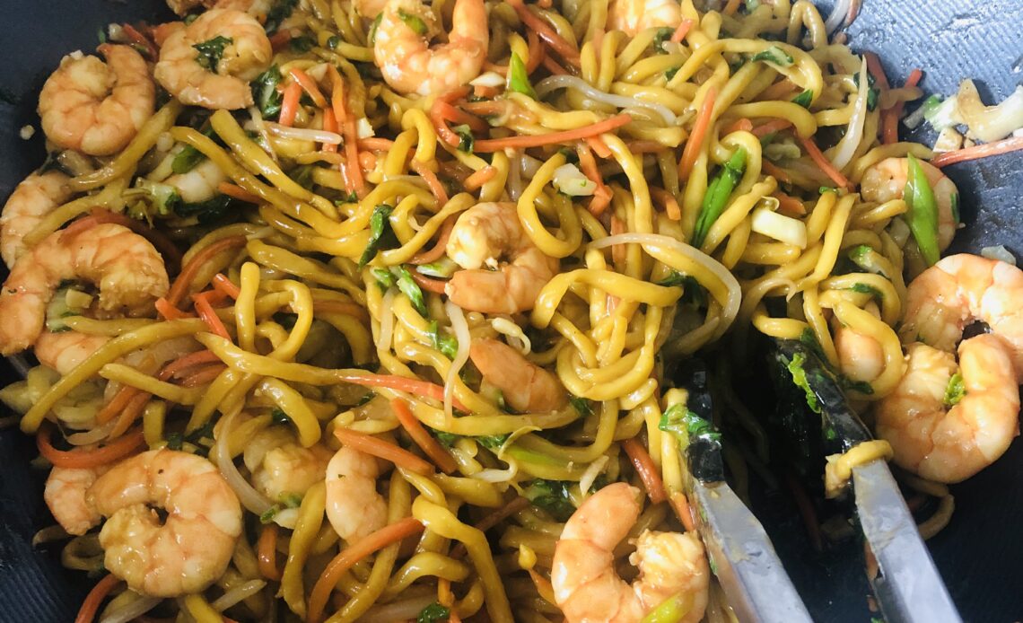 A mound of Chinese lo mein topped with shrimp and veggies in a large non-stick wok