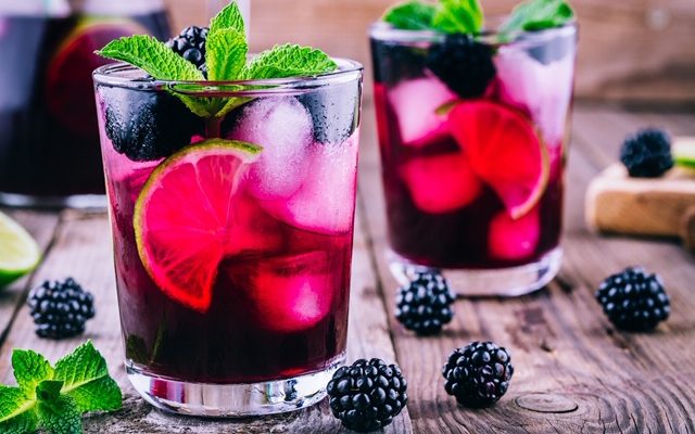 Two glasses sitting on a wooden farm table that has scattered blackberries and mint leaves on it. The glasses contain freshly made blackberry mojitos with large chunks of ice, half lime slices, blackberries and mint on top.