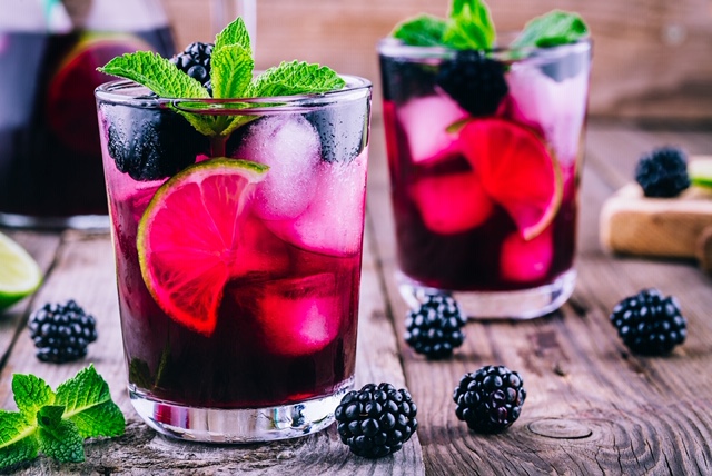 Two glasses sitting on a wooden farm table that has scattered blackberries and mint leaves on it. The glasses contain freshly made blackberry mojitos with large chunks of ice, half lime slices, blackberries and mint on top.