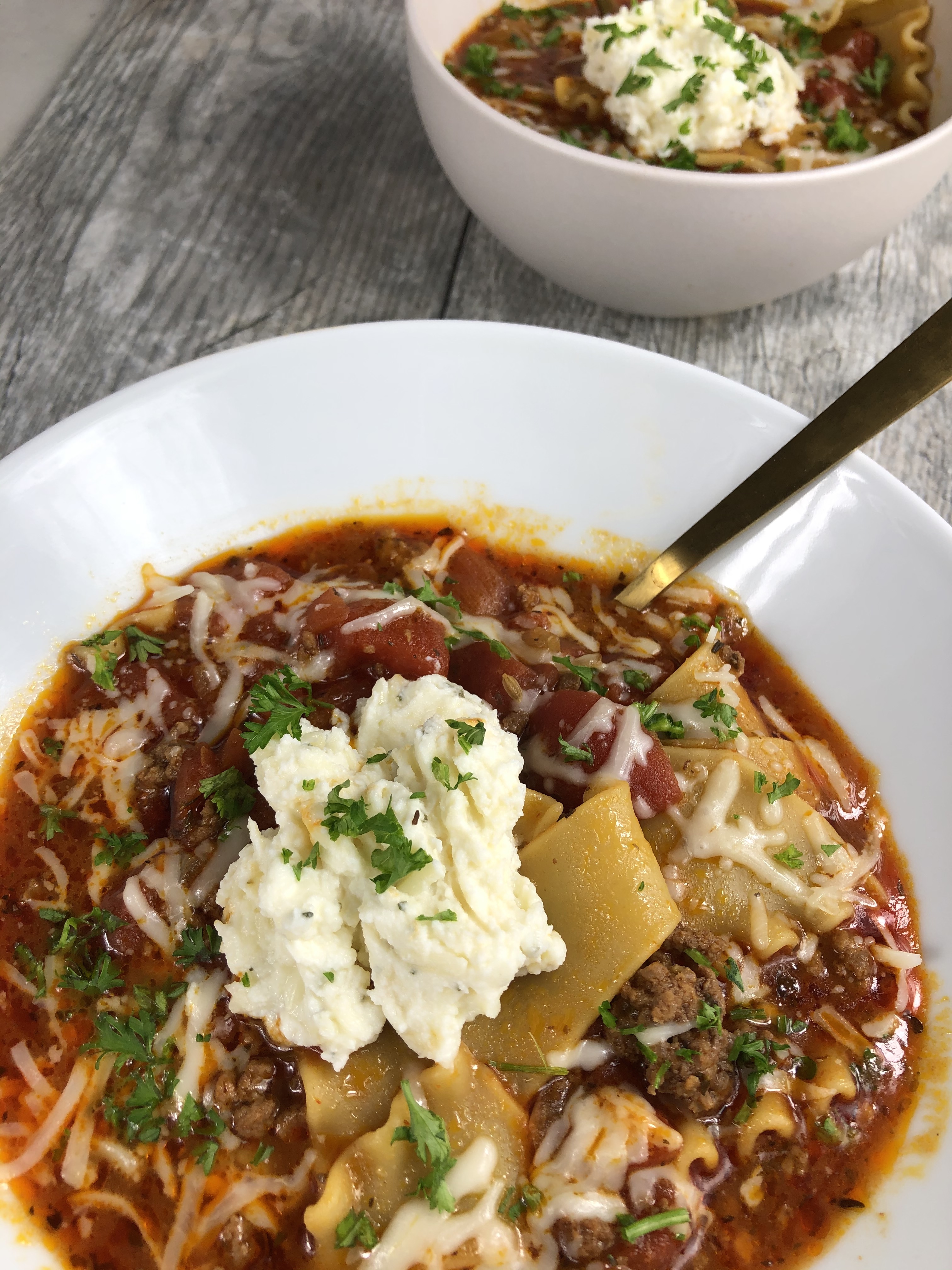 White porcelain bowl filled with lasagna soup. Pieces of ground beef and shreds of Parmesan cheese poke through the tomato sauce. A large dollop of ricotta cheese is on top of the soup in the center of the bowl and sprinkled with parsley leaves.