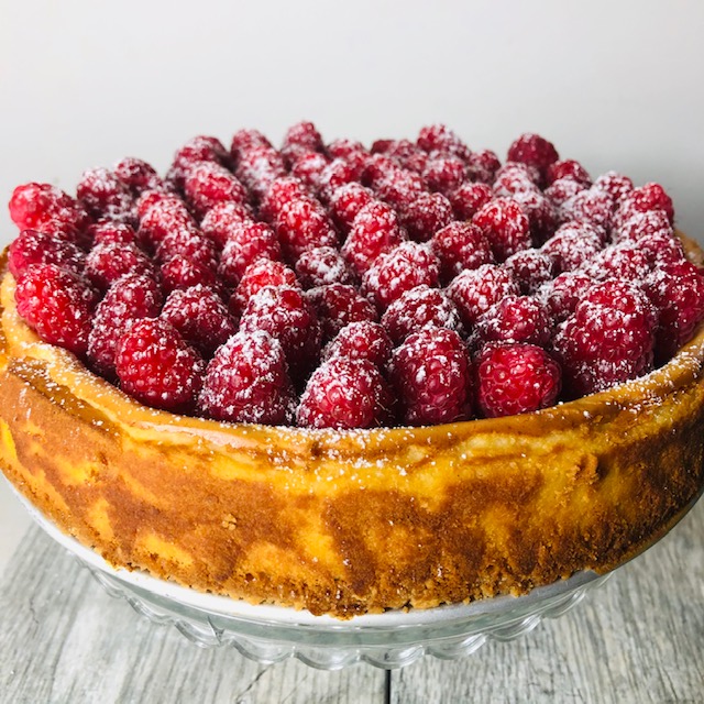 Shortbread crust cheesecake topped with raspberries and sprinkled with confectioner's sugar on crystal cake stand.
