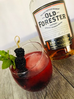Blackberry Bourbon Cocktail in a glass garnished with fresh mint and blackberries with a bottle of Old Forrester Bourbon in the backrgound.