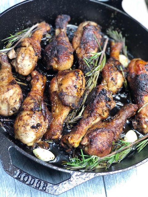Large cast iron skillet filled with crispy chicken drumsticks in a honey-soy and balsamic vinegar sauce and tossed in shallots, garlic, rosemary and black peppercorns.