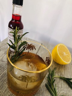 A gold-rimmed cocktail glass holding a compressed ice ball and filled with a boubon cocktail garneshed with lemon rind and a sprig of rosemary. In the background, there is half of a lemon and a bottle or aromatic bitters. In the foreground, a single sprig of rosemary lays on a flat wooden surface.