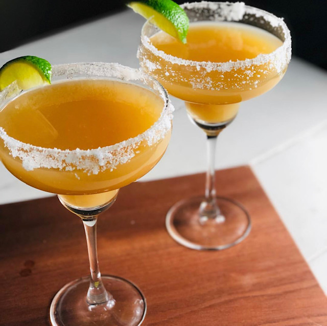 Two guava margaritas sit in margarita glasses with sugared rims and each garnished with a lime wedge on top of a dark wood plank against a white background.