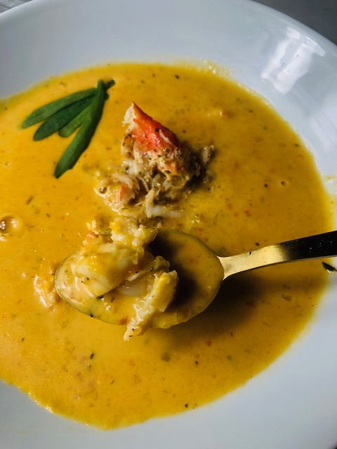 Lobster bisque topped with tarrogn leaves in an enamel white bowl with a golden soup spoon filled with bisque and a large chunk of seasoned lobster meat