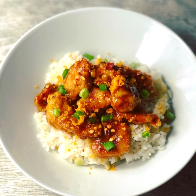 A white porcelain bowl filled with steamed white rice and topped with crispy orange chicken garnished with red pepper flakes and chopped scallion.