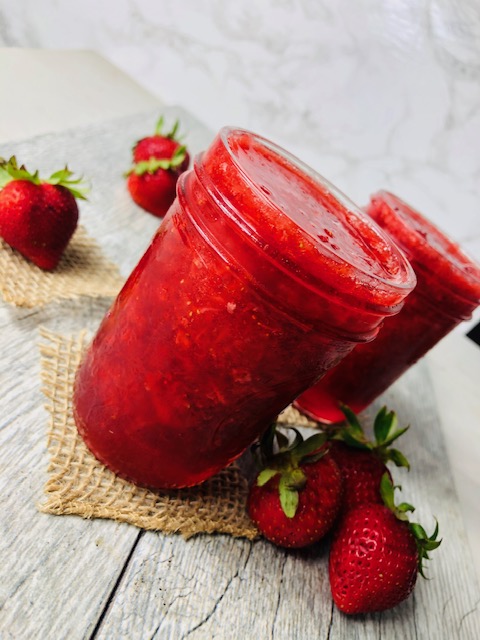 Strawberry Freezer Jam in open 8oz mason jars on slips of burlap surrounded by fresh strawberries on a wooden slab.