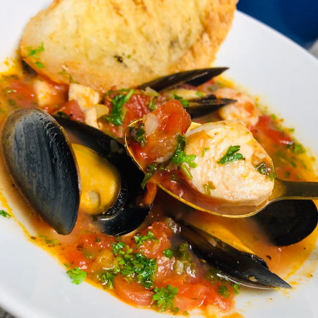 Mussels, mahi mahi, and shimp bathed in a wine and tomato stew and topped with fresh parsely served in a white enamel bowl with a slice of French baguette.