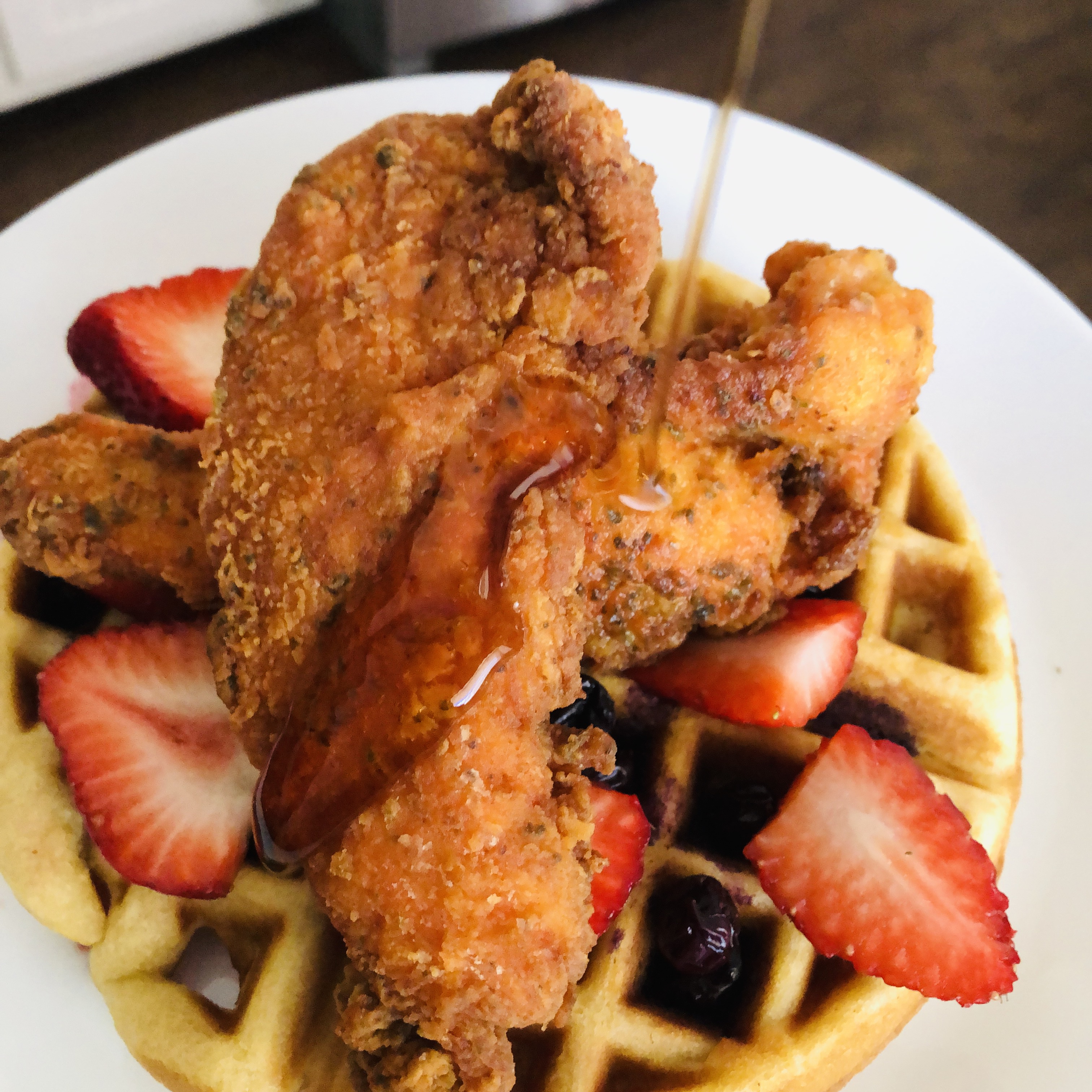 Belgian waffles topped with fresh strawberries and blueberries and fried chicken tenders with maple syrup being poured onto the stack.