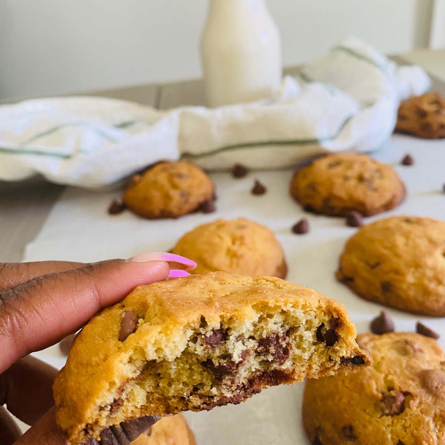 Chocolate Chip Cookies on a sheet of white parchment paper sprinkled with chocolate chips with a striped kitchen towel and a milk carafe in the background.