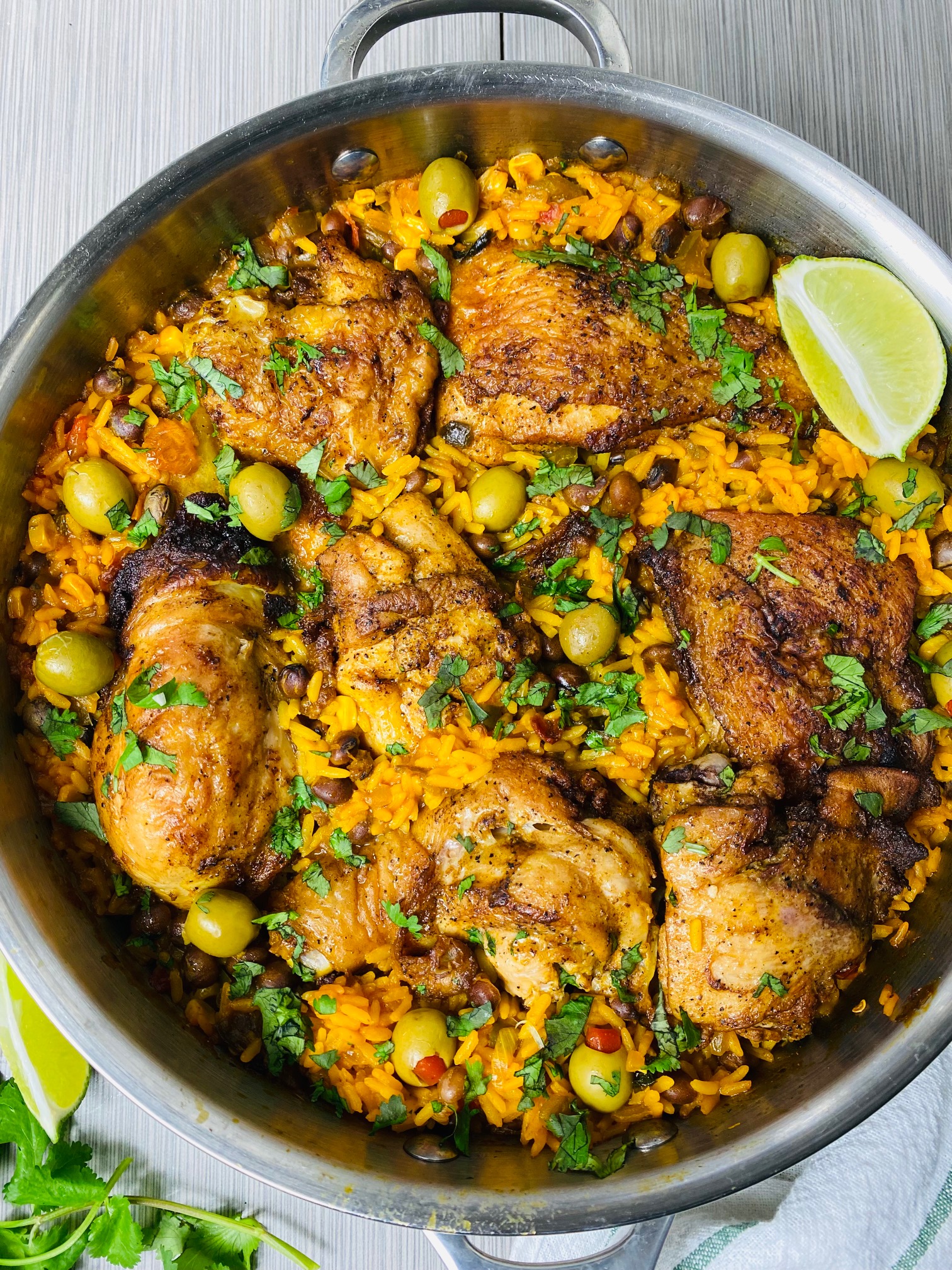 Seasoned, pan seared, crispy skin chicken served in a skillet with yellow rice and manzanilla olives and garnished with cilantro and a lime wedge served in a deep stainless steel skillet