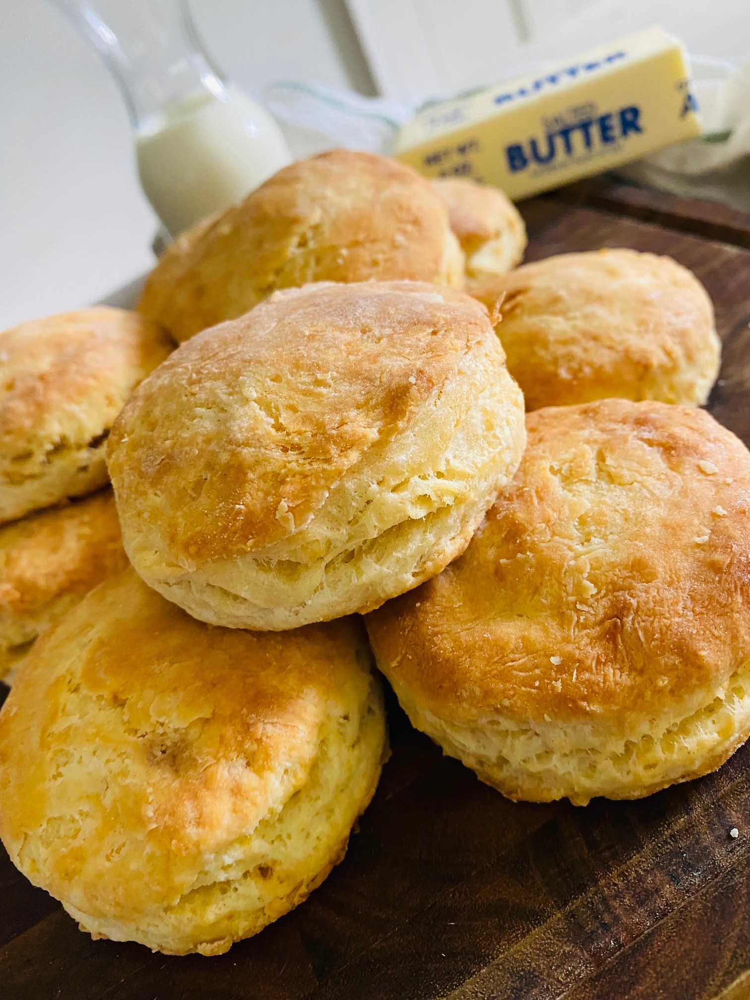 A pile of flaky, golden buttermilk biscuits on a dark colored wood curring board with a container of buttermilk and a stick of butter in the background.