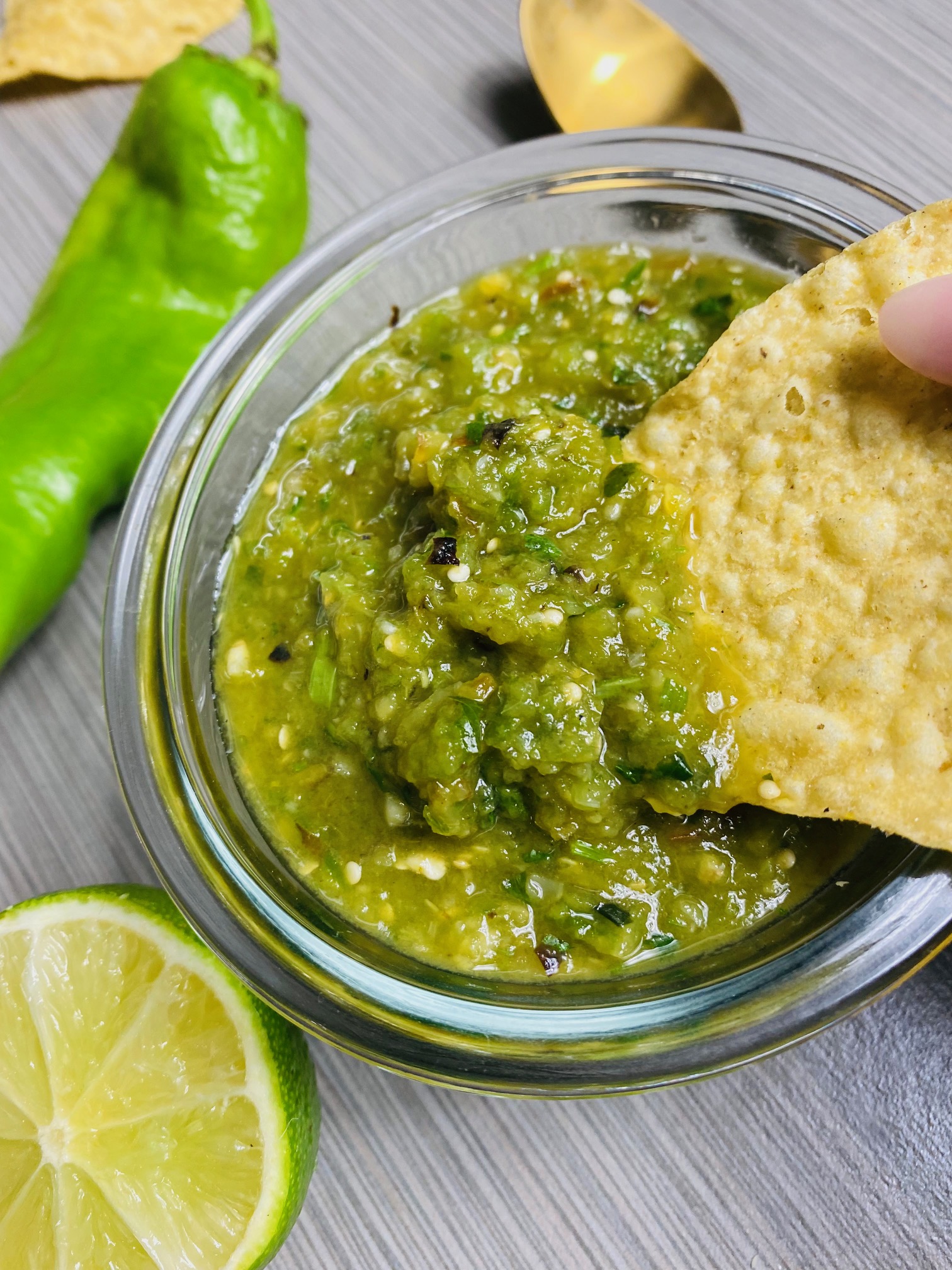 Salsa verde in a clear serving dish with a tortilla chip being dipped into the salsa. The serving dish is surrounded by a half of lime, a Hatch chile, a gold spoon, and more tortilla chips in the background.