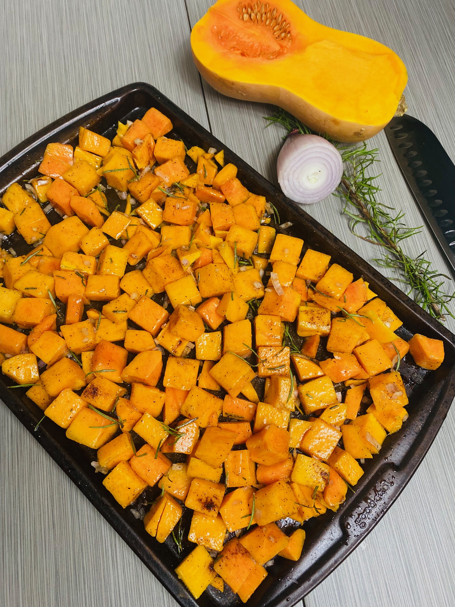 Chopped butternut squash seasoned with olive oil, rosemary, cinnamon, nutmeg, salt, pepper, and shallots on a nonstick baking sheet surrounded by half of a peeled shallot, a sprig of rosemary, half of a butternut squash and a chef's knife against gray tiles