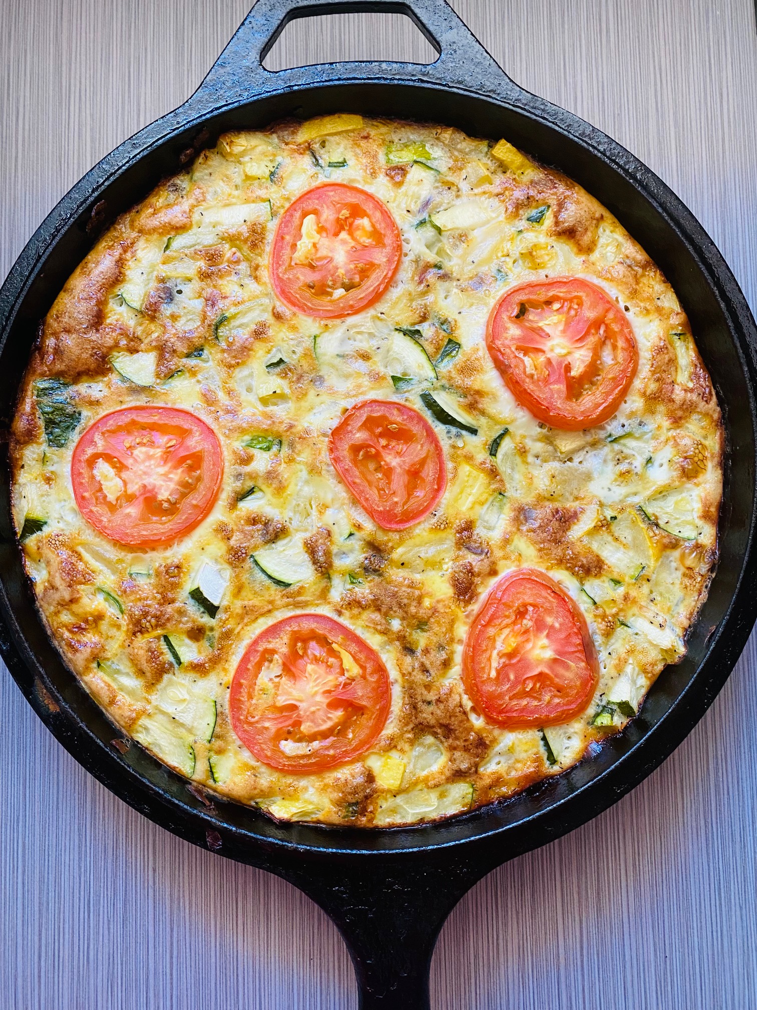 Veggie frittata made with zuchinni, yellow squash, red onion, and eggs, topped with tomato slices in a cast iron pan against a grey tile background.